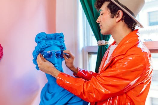 Stylish man in orange jacket and hat puts sunglasses on a vibrant blue bust indoors.